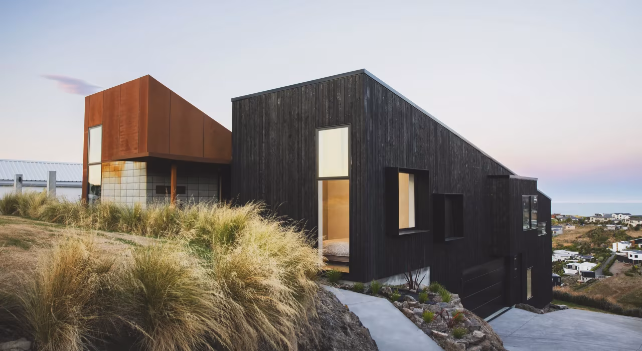 <img alt="Drayton exterior at dusk, charred larch and coretn steel volumes set among tussock grasses on the hillcrest." src="/images/s/i/t/e/-/site-architecture-drayton-drive8-39874007.webp" width="2560" height="1706" />